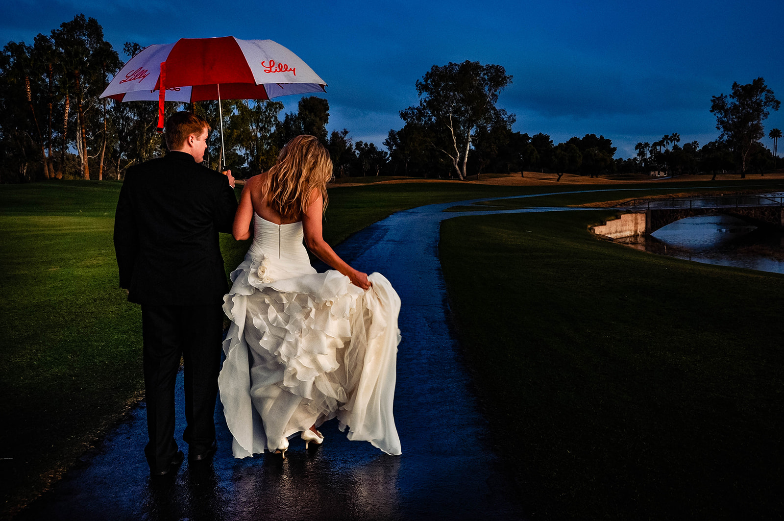 Bride and Groom walking on a wet golf cart path through a golf course next to a lake at the McCormick Ranch Golf Club on a dark, gloomy rainy day in Scottsdale, Arizona. The clouds are dark blue and grey. The bride is holding up her white wedding dress and she walks next to the groom who is in his black tuxedo carrying a red and white pattern umbrella over his and her head.