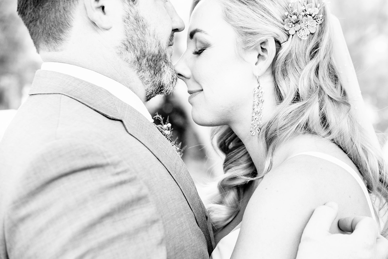 Black and white photo taken at a private residence in Paradise Valley, Arizona. Close up of a bride and groom in an intimate moment. Bride has her eyes closed, her nose is resting on the groom’s chin during the first look. His right hand is caressing her back. She has long diamond earrings on and a floral hair piece on the left side of her hair.