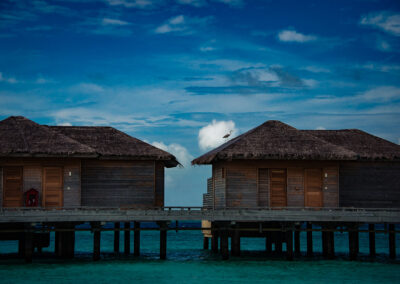 Two hut-like over water villas in the Maldives at the You and Me Cocoon Resort. There is a grey heron on top of the villa on the right of the photo. It is framed in the white cloud in the distance, while the rest of the sky and the water is blue.
