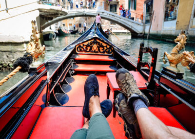 Two feet on the seats in a gondola in Venice, Italy. The gondola is red and black with gold lining and gold horse statues.